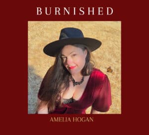 Burnished CD cover, burgundy frame inner image of Amelia in wide brimmed black hat with painted flowers in red cress seated on Golden dry grassy ground.